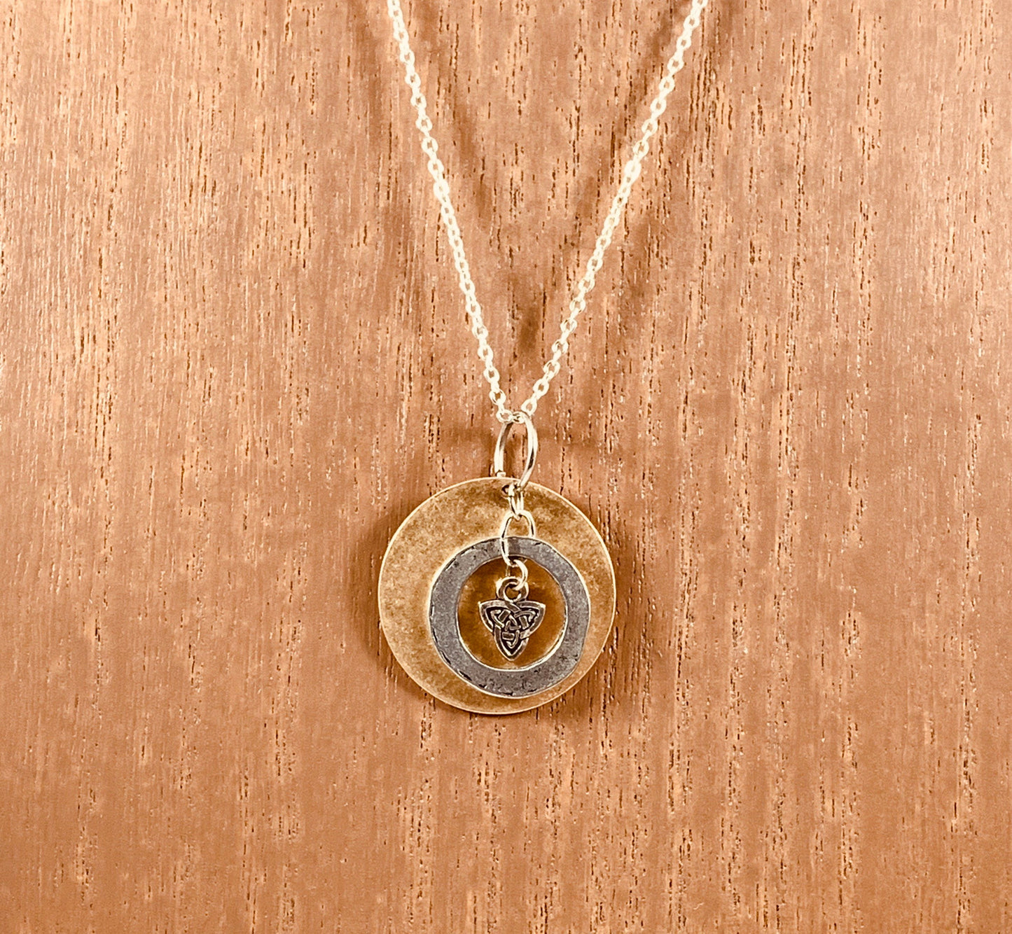 Sterling silver Celtic knot encircled with silver ring and placed in front of brass disc suspended from bronze colored chain necklace.